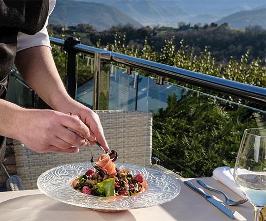 Imagen de un camarero sirviendo un plato de comida de autor en una terraza con el paisaje de fondo