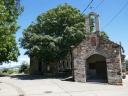 Pequeña ermita de piedra con campana en el centro situada junto a un gran árbol frondoso