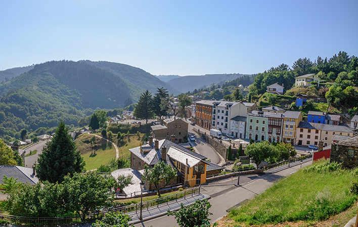 Blick auf Santalla, den Hauptort von Santa Eulalia de Oscos, ein malerisches asturisches Dorf. Bunte Häuser schmiegen sich an die Hänge und bilden ein Mosaik aus Fassaden, das mit der üppigen Vegetation kontrastiert. Im Hintergrund erhebt sich eine bergige Landschaft, während ein blauer Himmel mit weißen Wolken der Szene Leuchtkraft verleiht.