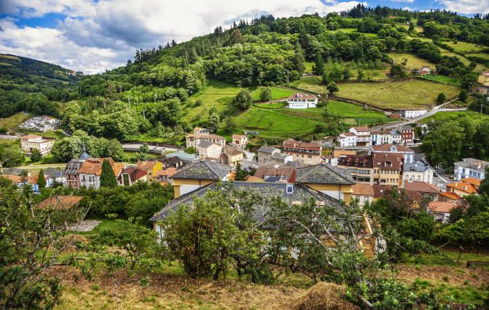 Vista de Pola de Allande, casas de diferentes estilos con tejados negros de pizarra, tradicional en esta zona y otras con tejados rojos. Detrás del pueblo se eleva una suave montaña salpicada de bosques y alguna casa más alejada. En primer plano una casona con finca y hórreo a la izquierda.
