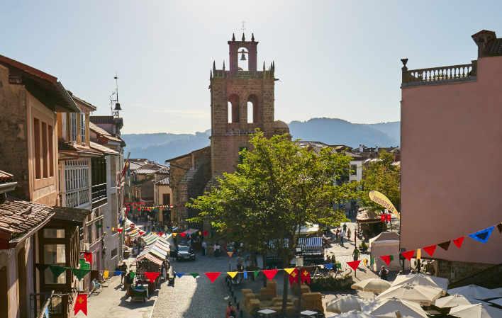 Veduta di Salas, in primo piano le bandierine colorate che decorano il villaggio in occasione della sua festa medievale, su entrambi i lati della foto vecchie case con ballatoi in legno che si affacciano sulle strade acciottolate e sulle piazze piene di bancarelle di prodotti tipici. Al centro si erge la chiesa con il suo campanile, con le montagne sullo sfondo.