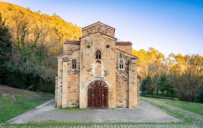 Vista exterior da igreja de San Miguel de Lillo sob um céu azul claro, mostrando a cantaria e o desenho arquitetónico. Por detrás do edifício vêem-se as árvores circundantes.