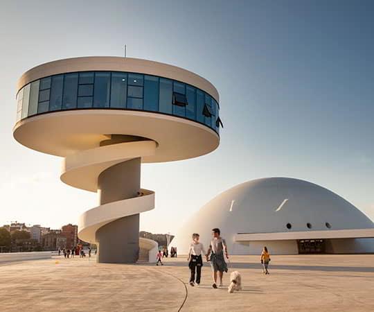 Ein Paar geht mit seinem Hund auf dem Niemeyer-Platz in Aviles spazieren, mit dem Turm und der Kuppel im Hintergrund.
