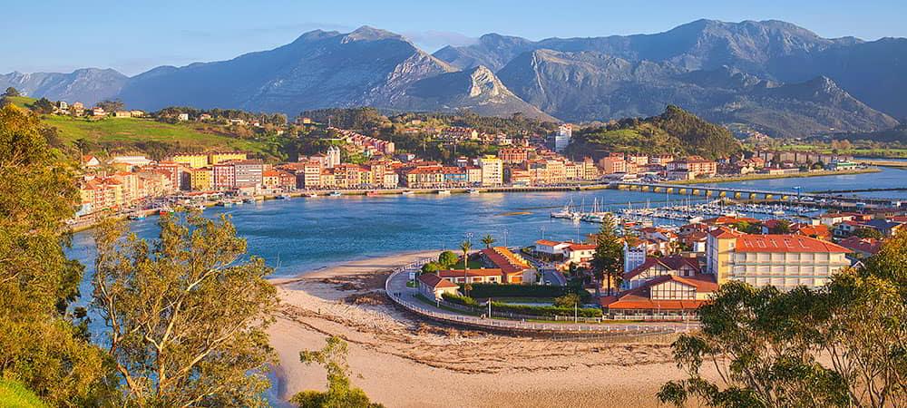 Vista panorámica del pueblo marinero de Ribadesella/Ribeseya en el que se ven el puerto y las casas del pueblo con las montañas al fondo.