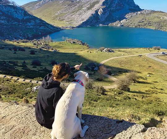 Paisaje montañoso asturiano con una mujer señalando el Lago Enol mientras su perro la acompaña, disfrutando de las vistas en un día soleado.