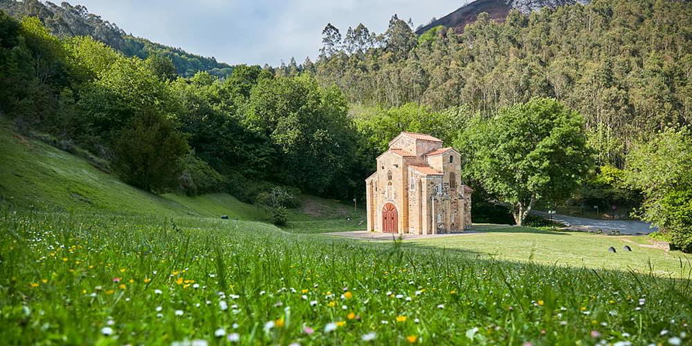 Vista panorâmica de San Miguel de Lillo, com a igreja a destacar-se no meio de um campo de erva alta com flores silvestres e árvores ao fundo.