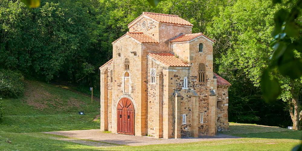 A fachada emblemática da igreja de San Miguel de Lillo, nas Astúrias, destacando a sua arquitetura única e rodeada de vegetação.