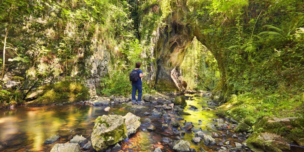 Mujer de espaldas en una roca en el río Tuernes, en Los Covarones de Tuernes (Llanera), vestida con vaqueros, camiseta azul marino y mochila negra; se encuentra frente a un arco natural de roca caliza cubierto de vegetación, con el riachuelo fluyendo por debajo y vegetación ribereña alrededor.