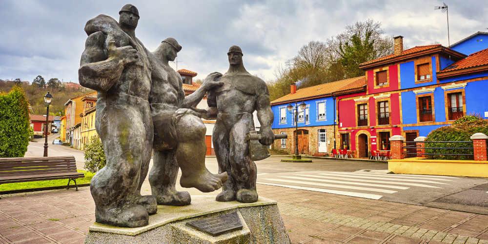 Monumento al minero en Carbayín Bajo (Siero): tres figuras macizas en bronce que representan mineros; uno sostiene un hacha en la mano izquierda, otro carga un bulto sobre el hombro y la figura central se apoya en los compañeros mientras levanta la pierna derecha en actitud de esfuerzo. Las esculturas, de apariencia robusta tipo “Hulk” o “El Hombre de Piedra”, se alzan sobre un pedestal de piedra. Al fondo, tras un paso de peatones, se distinguen casas de vivos colores granate y azul en un entorno urbano abierto.