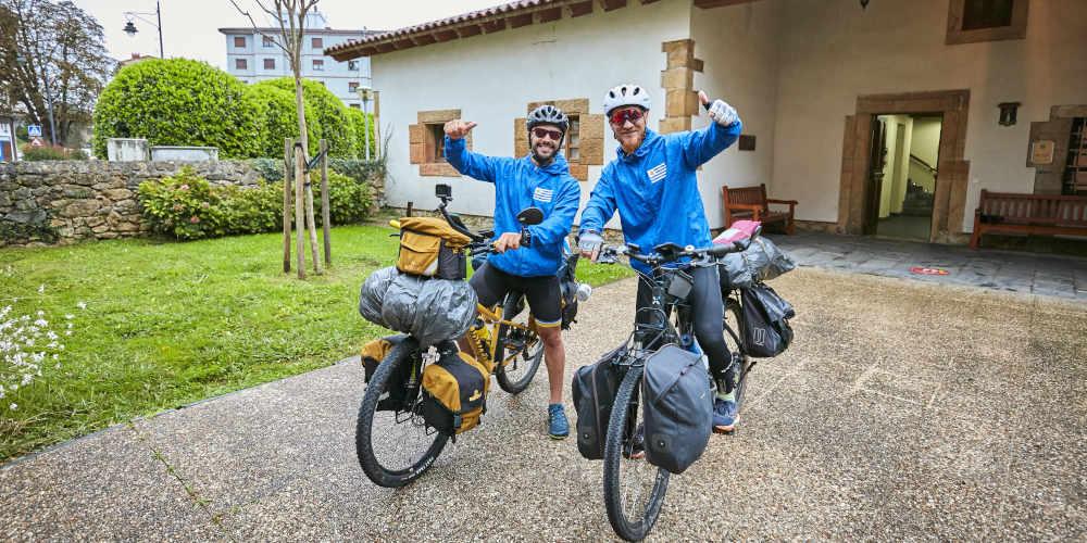 Dos peregrinos en bicicleta sobre el Camino de Santiago, equipados con mochilas y bultos atados a las bicicletas en la parte delantera y trasera; ambos visten chubasqueros azul cielo con una bandera similar a la argentina, llevan gafas ciclistas y posan sonrientes levantando los pulgares, junto a un paso empedrado con un pequeño jardín y un árbol al lado.