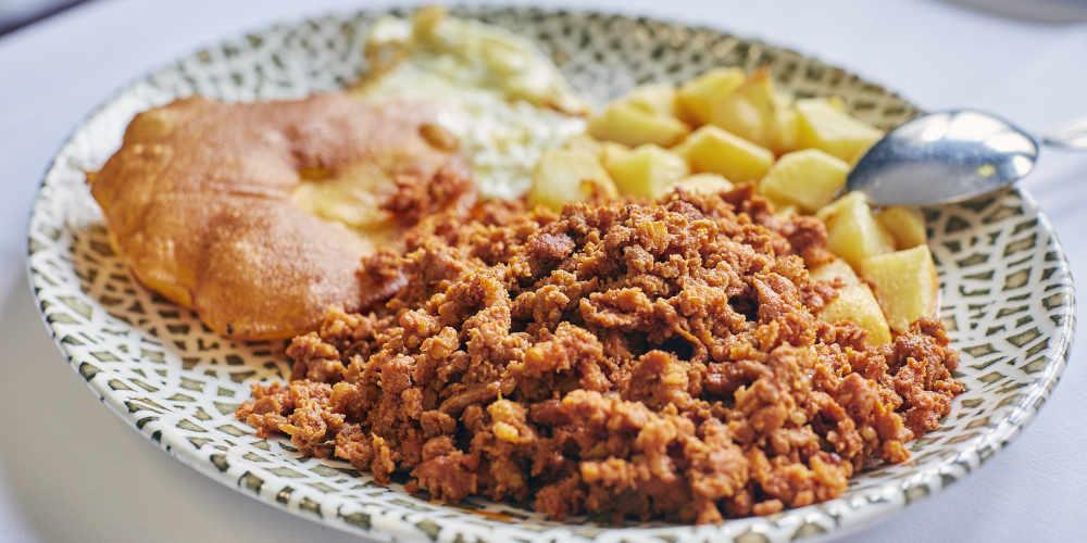 Primerísimo plano de un plato de picadillo con tortos y patatas, servido en un plato decorado con motivo de mosaico sobre mantel blanco, acompañado de una cuchara apoyada en el borde.
