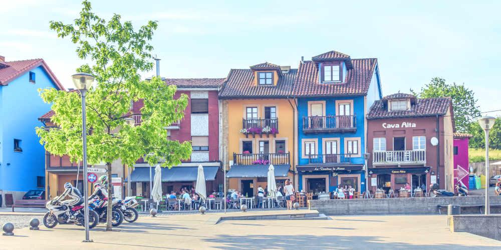 Plaza Les Campes en Siero, con casas de fachadas pintadas en colores intensos amarillo, azul y marrón; bajo los soportales se ven terrazas con gente tomando algo, mientras en la plaza destacan un árbol y varias motos estacionadas junto a la zona pavimentada despejada.