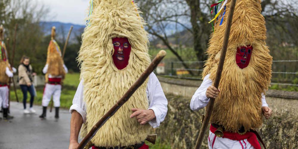 Dos Sidros en primer plano durante la mascarada de invierno de Valdesoto, con el traje tradicional formado por camisa y pantalón blancos, faja y cintas de colores, careta y un largo palo en la mano; al fondo, difuminados, se distinguen otros dos Sidros de espaldas.