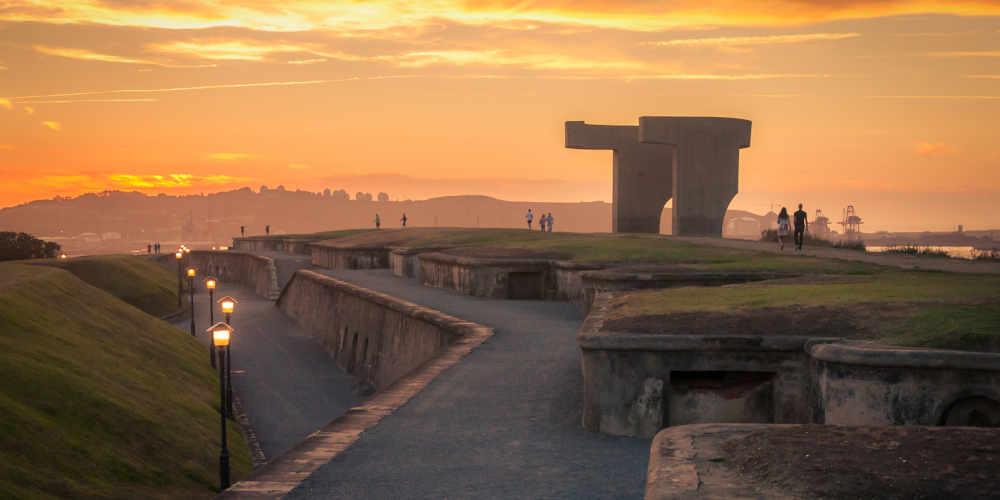 Monumento Elogio del Horizonte en la parte alta de Cimavilla, fotografiado de lado durante un atardecer de tonos naranjas intensos; alrededor se observan paseantes, farolas encendidas en la zona inferior y, al fondo, el paisaje urbano de Gijón con referencias como las instalaciones industriales y las bombonas de la Campa Torres.