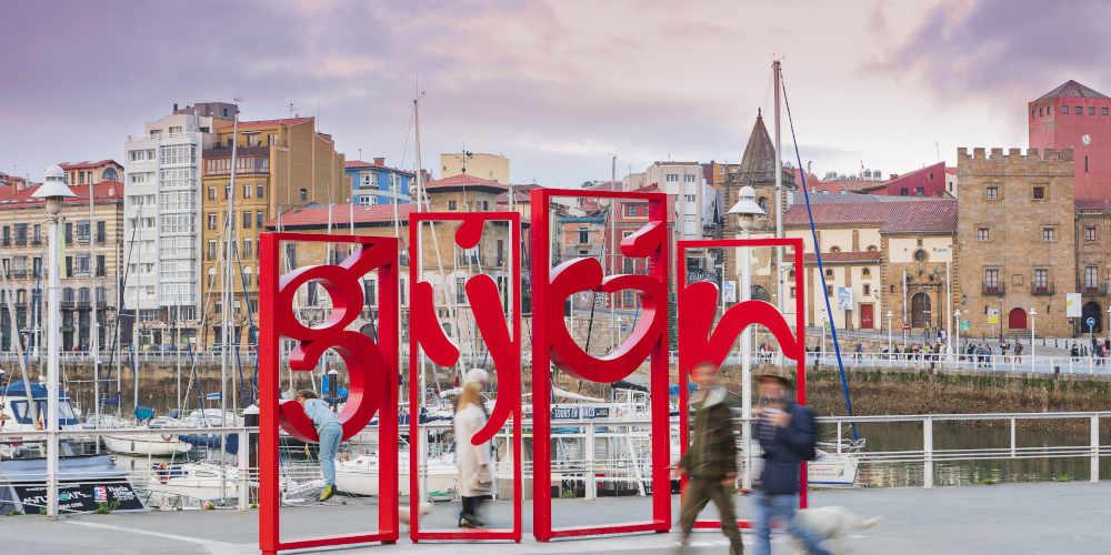 Letrero rojo de las letronas de Gijón con paseantes en primer plano; detrás, el puerto deportivo con barcos y veleros, edificios del centro, la Torre del Reloj y el Palacio de Revillagigedo.