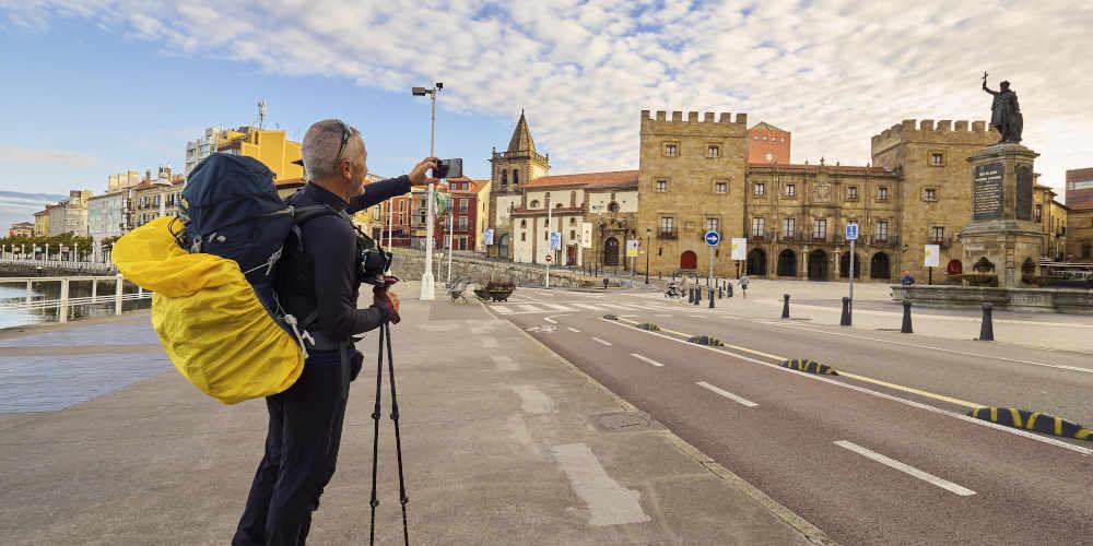 Peregrino en el paseo del puerto deportivo de Gijón tomando una fotografía con su teléfono móvil hacia la estatua de Pelayo y el Palacio de Revillagigedo; lleva mochila de caminante y sostiene en la mano los palos de senderismo.