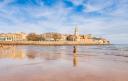 Panorámica de la playa de San Lorenzo en Gijón con la ciudad al fondo y una mujer paseando por la orilla