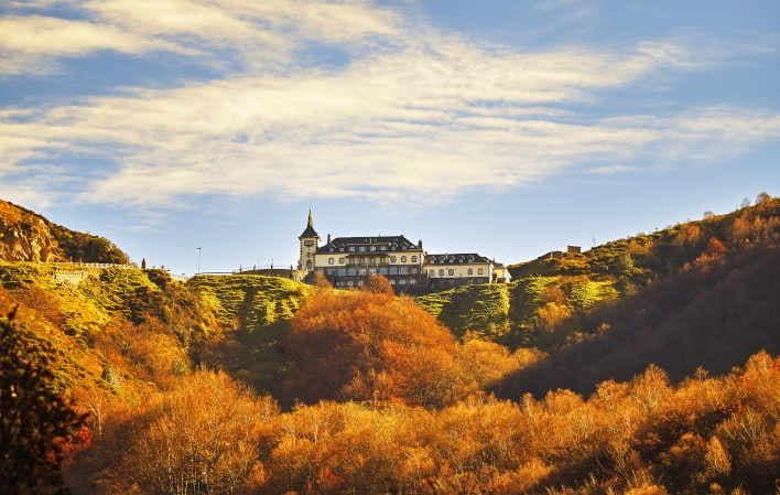 Antiguo Parador de Pajares en Lena, rodeado de un entorno natural en tonos tierra anaranjados, con el edificio al fondo bajo un cielo parcialmente despejado.