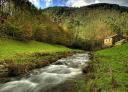 Río de aguas rápidas entre praderas y bosque con cabaña de piedra