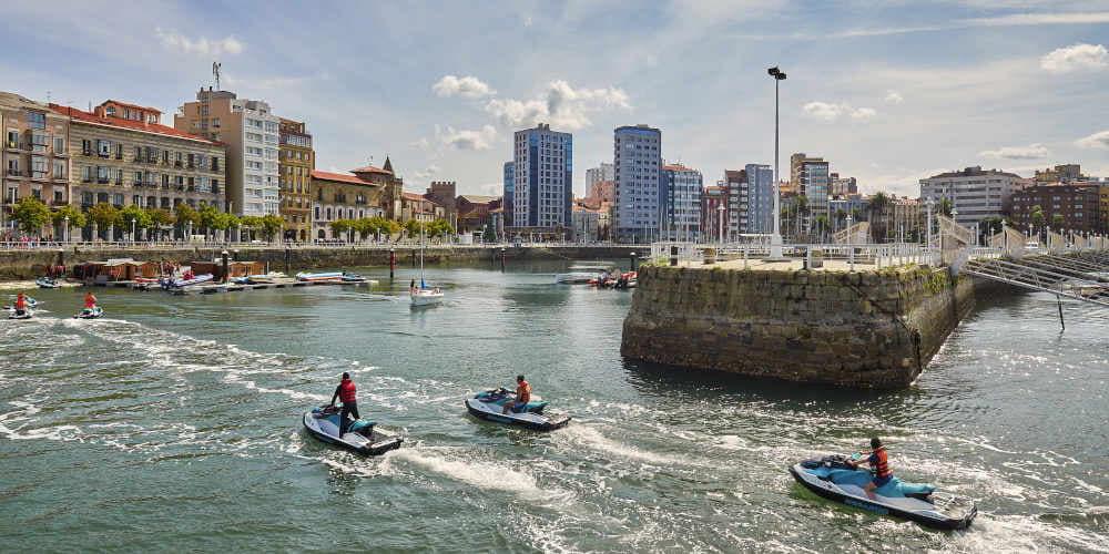 Zona de alquiler de motos de agua en el puerto deportivo de Gijón, con tres personas regresando en sus motos hacia el muelle; al fondo se distingue el paseo del puerto, el monumento de las botellas de sidra y varios edificios urbanos.