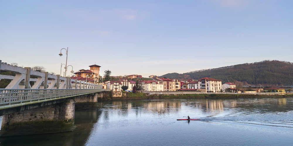 Ría Tina Mayor en Bustio, vista en amplia extensión con un único piraguista avanzando hacia un puente situado a la izquierda; al fondo se distinguen edificios junto a la ribera.