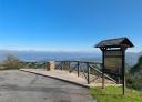Miradouro com vedação de madeira e placa informativa à direita com vista para as montanhas.