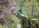 Hiker walking through a narrow pass with a stone wall and vegetation.