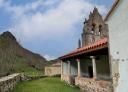 Stone church with a bell tower in a green setting next to a low stone wall.