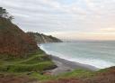 View from the top of a beach with dark sand, green and reddish cliffs and calm sea.