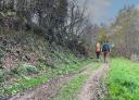 Couple of hikers on a dirt and grassy path surrounded by vegetation.