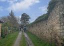 Dos personas caminan junto a un muro de piedra en un sendero rural