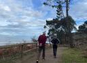 Dos personas caminando por un sendero con barandilla de madera en la costa