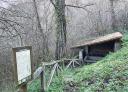 Old wooden and stone washhouse next to an information panel, surrounded by vegetation.
