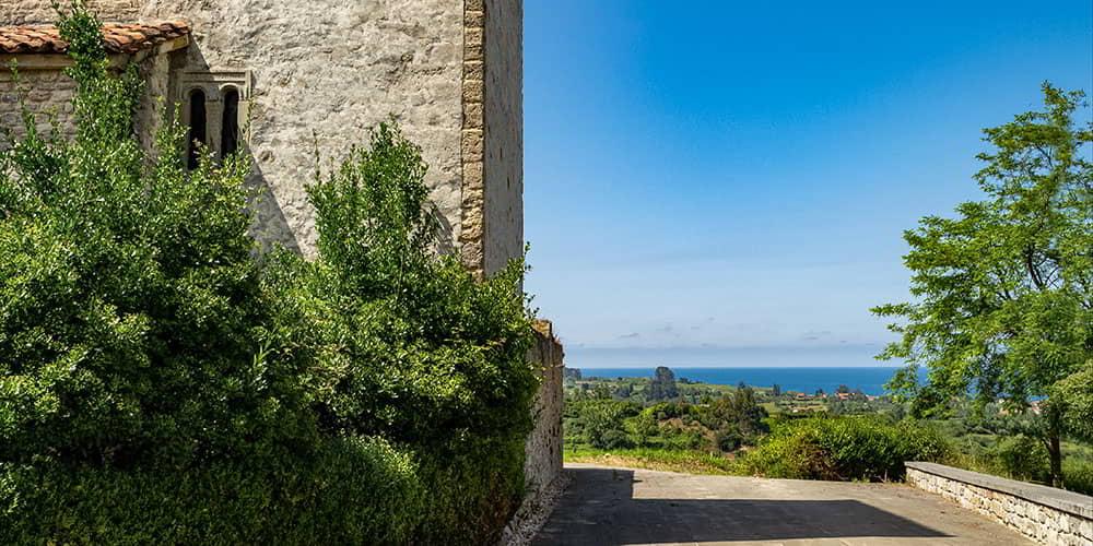 Lado da igreja de Santiago de Gobiendes, mostrando uma janela pré-românica e a paisagem asturiana com o mar ao fundo.