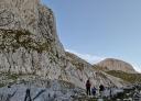 Hikers walk through a mountain valley, surrounded by towering rocky peaks and blue skies.