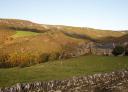 Rural valley with green fields and stone house, surrounded by wooded hills.