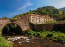 Antiguo monasterio de piedra con arcos, junto a un río y puente histórico en un entorno verde