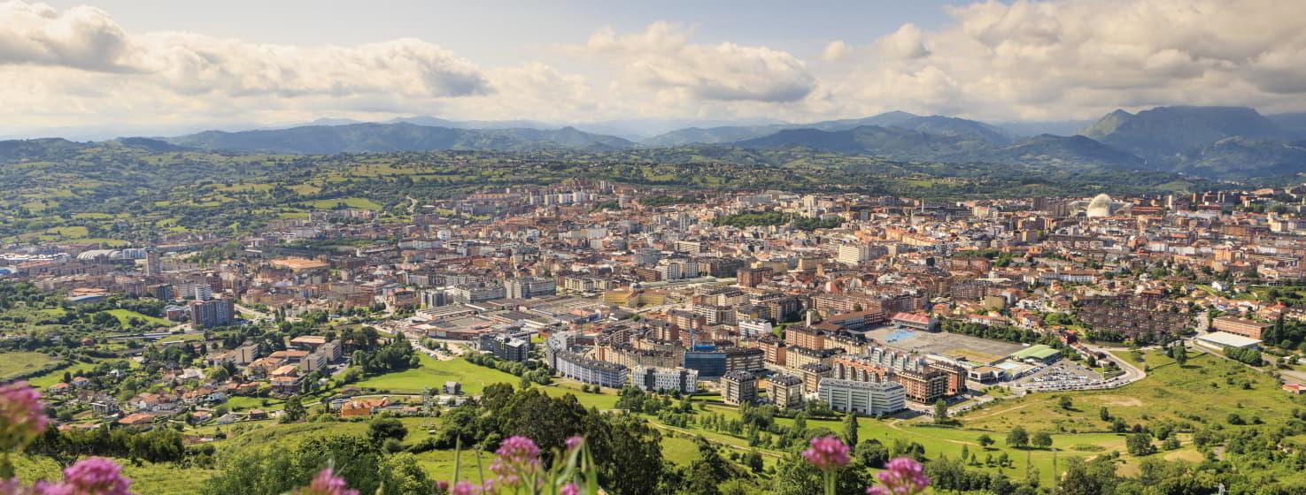 Vista panorámica de la ciudad de Oviedo desde el monte Naranco, con la trama urbana extendiéndose entre prados y montañas, bajo un cielo parcialmente nublado.