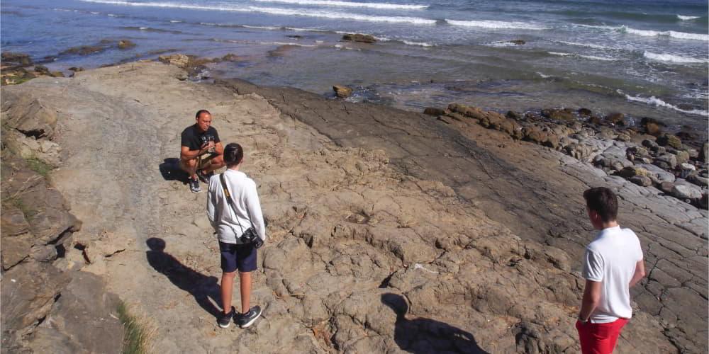 Yacimiento de icnitas en la playa de La Griega, en Colunga, con tres personas observando las huellas fósiles sobre la roca; una de ellas aparece agachada mientras las otras dos, de pie y de espaldas a la cámara, contemplan el yacimiento con la orilla del mar al fondo.