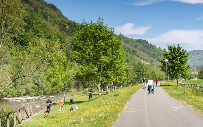 Senda fluvial del Nalón en Laviana, con caminantes y ciclistas recorriendo el camino asfaltado junto al río, rodeado de prados y árboles, en un entorno de montañas verdes bajo cielo despejado