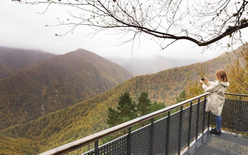 Mädchen mit Mackintosh am Aussichtspunkt Muniellos, das die Landschaft fotografiert, während es sich hinter einem perforierten Metallgeländer auf eine Fläche stützt; im Hintergrund ist eine wolkenverhangene und neblige Umgebung zu sehen, mit Tälern und Bergen, die mit grüner und ockerfarbener Vegetation bedeckt sind.