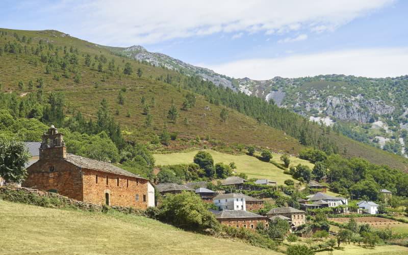 Blick auf das Dorf Seroiro in Ibias, mit einer Steinkirche im Vordergrund und mehreren traditionellen Häusern, die von Wiesen und Bäumen umgeben sind, am Fuße der mit Vegetation und Felsformationen bedeckten Berge unter einem teilweise wolkenlosen Himmel.