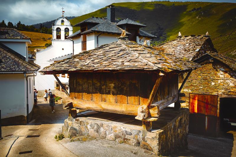 Ansicht von Besullo in Cangas del Narcea mit einem steinernen Getreidespeicher im Vordergrund neben einer abschüssigen gepflasterten Straße; hinter dem Getreidespeicher ein weißes Haus mit grauem Schieferdach und im Hintergrund eine weiße Kirche mit einem zweibogigen Glockenturm. Rechts, unterhalb der abschüssigen Straße, steht ein lagerhausähnliches Steingebäude mit einer großen Holztür, in der ein Auto steht. Das Ganze wird von grün bewachsenen Bergen unter einem wolkenverhangenen Himmel eingerahmt.