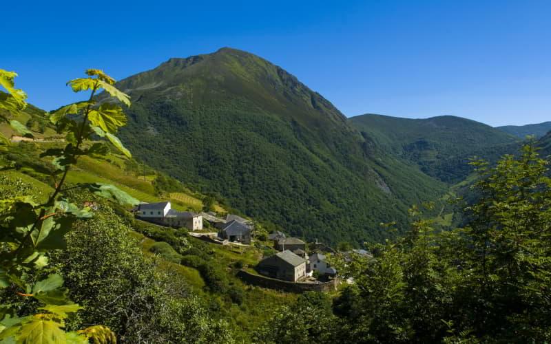 Ansicht von Brañas de Arriba von oben, mit einer Häusergruppe in der Mitte, umgeben von Wiesen und Bergen, und im Hintergrund der Cueto de Arbas unter einem völlig klaren blauen Himmel.