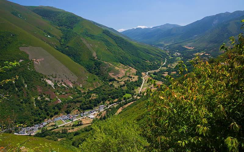 Panoramablick auf Fonduveigas in Degaña, mit dem Dorf in der Talsohle, umgeben von grünen Bergen und Hängen mit bewaldeten Flächen und Lichtungen, durchzogen von einer Straße, die sich unter klarem Himmel durch die Vegetation schlängelt.