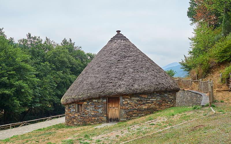 Ein Palloza in Ibias mit einem Steinsockel, einer Holztür und einem kleinen Fenster, überdacht von einem länglichen, kegelförmigen Strohdach; gelegen an einem Hang neben einer Baumgruppe, unter einem völlig bewölkten Himmel.