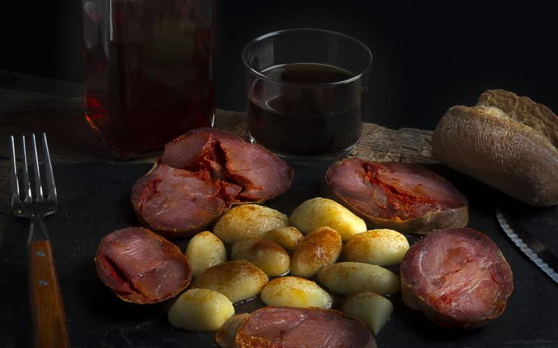 Gericht mit Chosco de Tineo und Salzkartoffeln, dazu Brot, ein Glas und ein Krug Rotwein auf einem rustikalen Tisch.