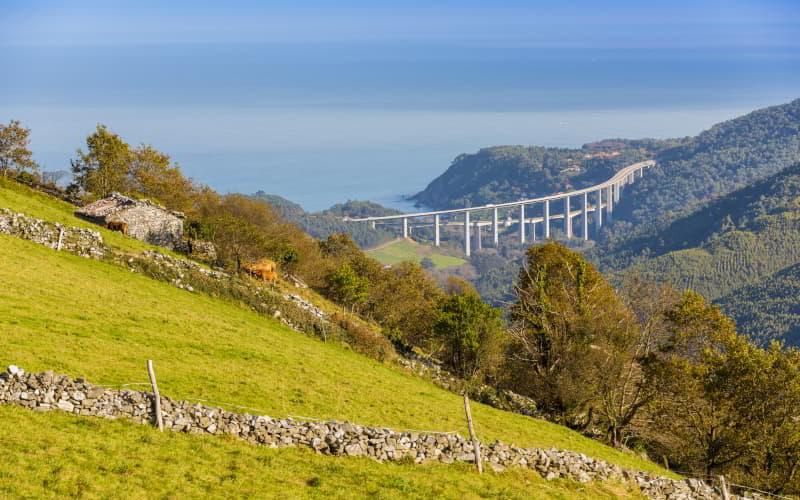Blick auf das Viadukt von Concha de Artedo von Brañaseca aus, mit grünen Wiesen und einer Steinhütte im Vordergrund, bewaldeten Hügeln ringsherum und dem Meer im Hintergrund.
