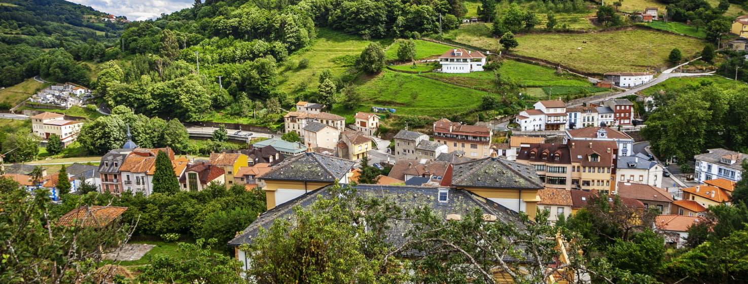 Blick auf Pola de Allande, Häuser verschiedener Stilrichtungen mit schwarzen Schieferdächern, wie sie in dieser Gegend üblich sind, und andere mit roten Dächern. Hinter dem Dorf erhebt sich ein sanfter Berg mit Wäldern und einigen weiter entfernten Häusern. Im Vordergrund ein großes Haus mit einem Bauernhof und einem Getreidespeicher auf der linken Seite.