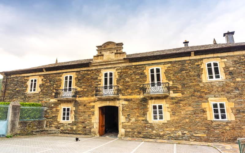 Fachada principal del Palacio de Ron, edificio de piedra con balcones de hierro forjado y portalón central en arco. El conjunto presenta elementos decorativos sobre la entrada y tejado de pizarra, con un espacio abierto pavimentado en primer plano.